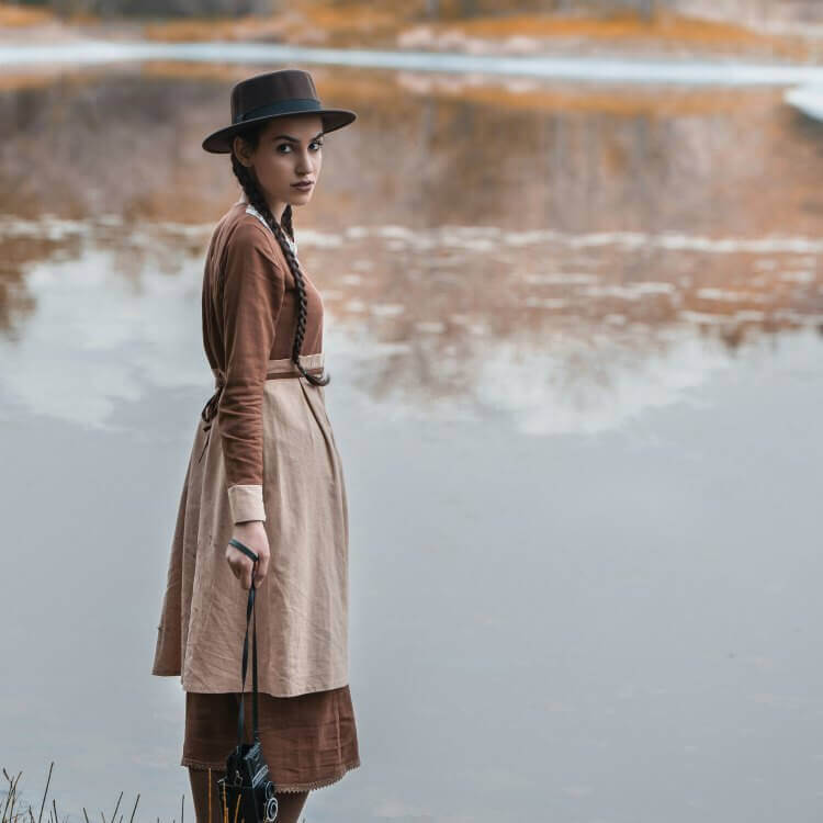 An image of a woman standing in front of a pond holding a film camera from its strap whilst looking towards the camera. She wears natural beiges and browns matching the colours behind her.