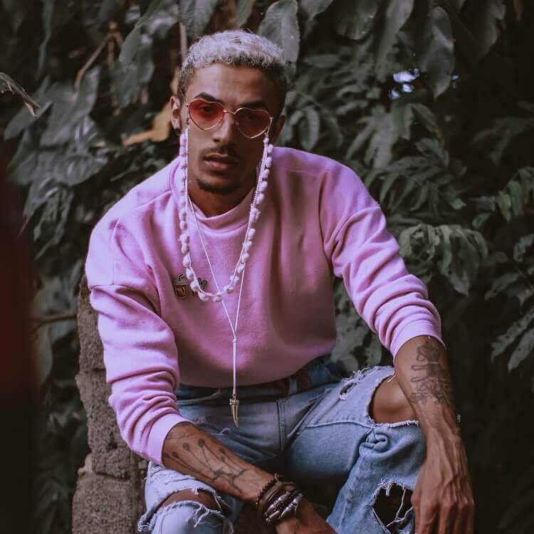 A photo of a man looking front on at the camera. He is sat in front of a plant filled wall wearing heart shades, a pink jumper and ripped jeans. 