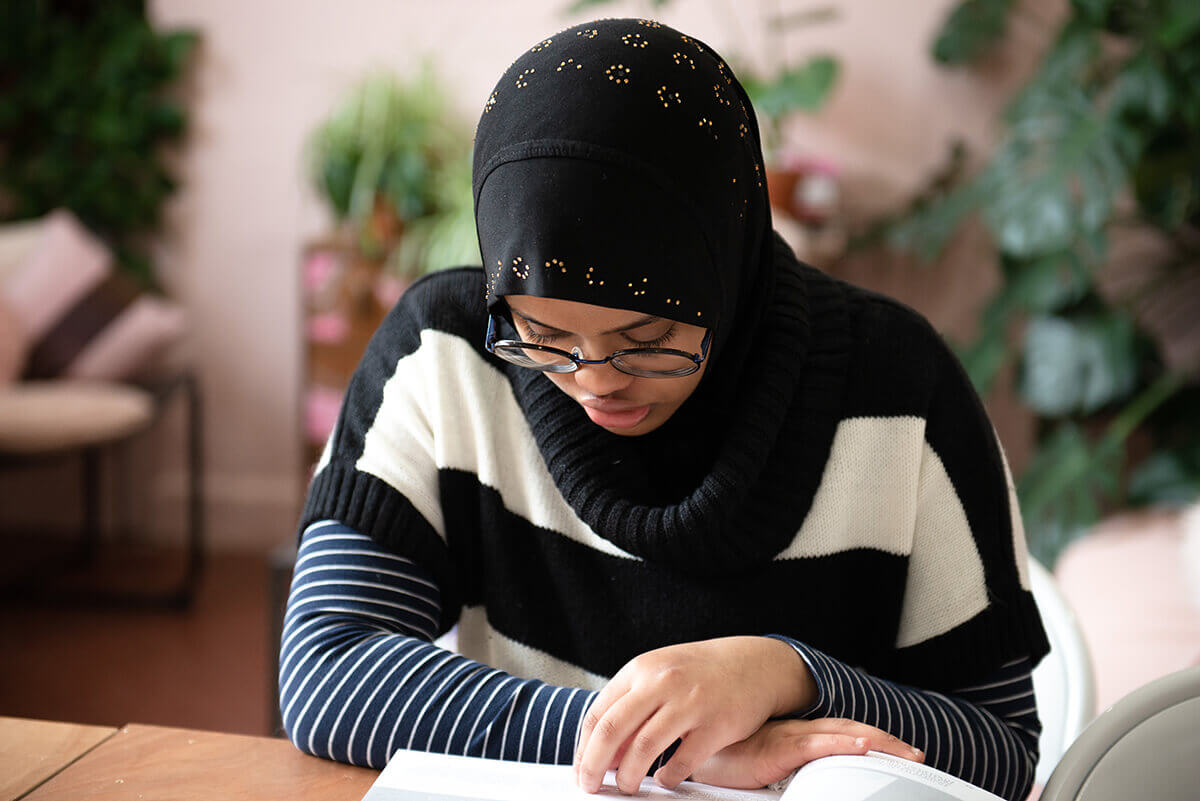 Work experience student Rayhan sits at a wooden desk, looking through a book. She is wearing a long sleeve striped tshirt underneath a black and white striped top.