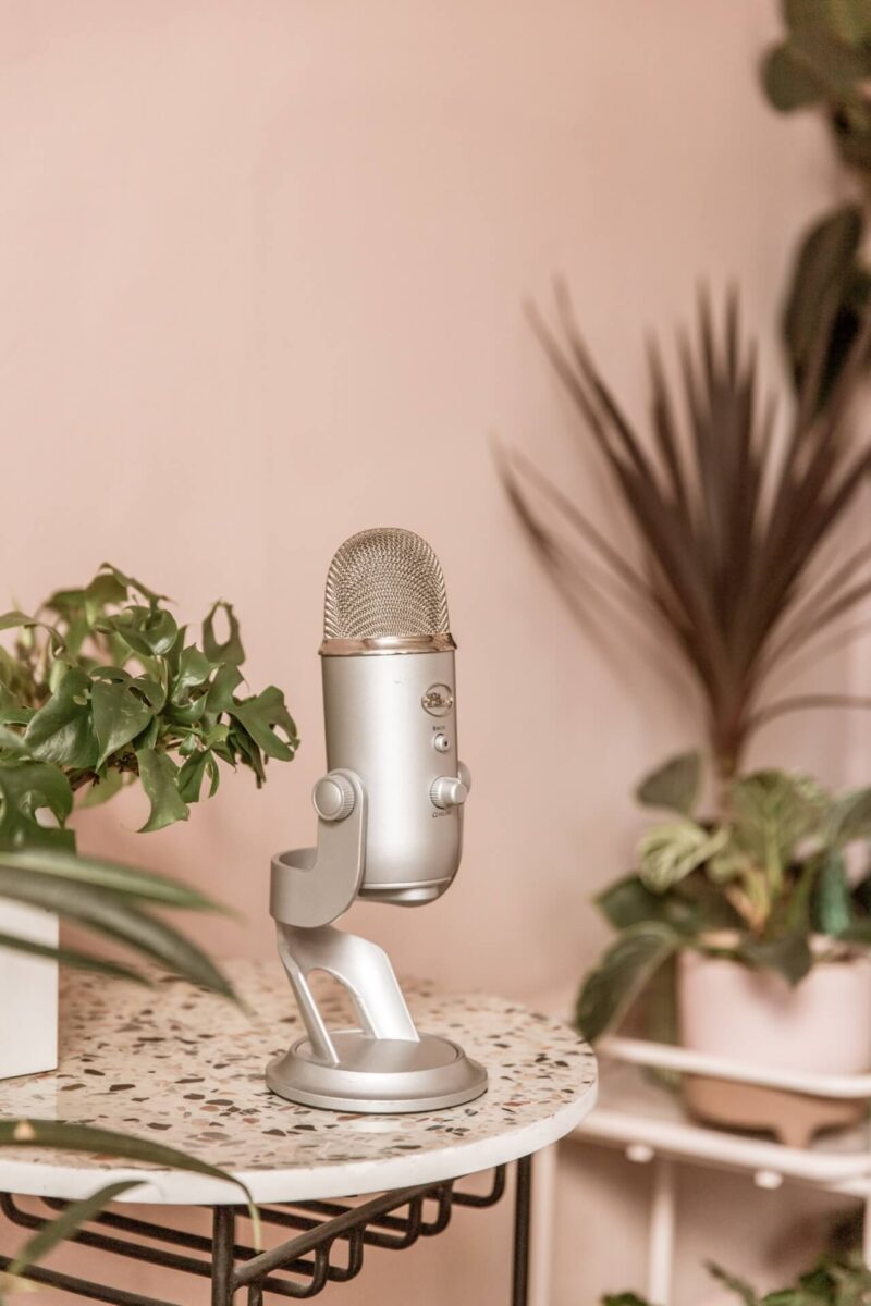 a podcast microphone sits on a terrazzo side table with Studio Cotton's pink walls as the back drop. It is surrounded by green plants.