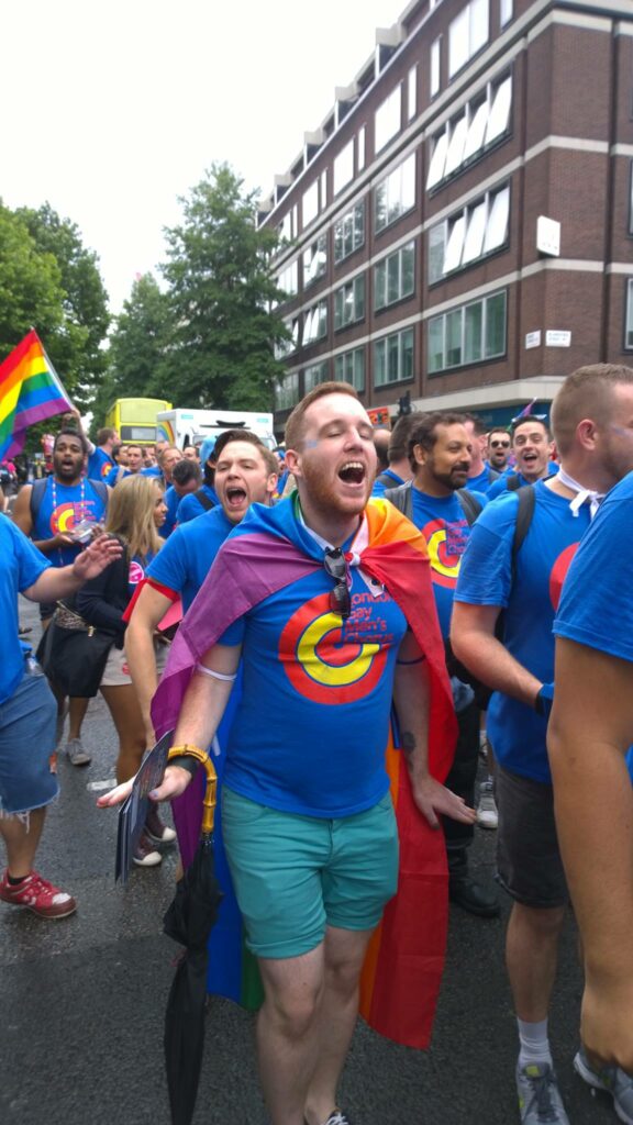 Adam at his first Pride, draped in a rainbow flag and colourful clothes, singing with the London Gay Men's Chorus