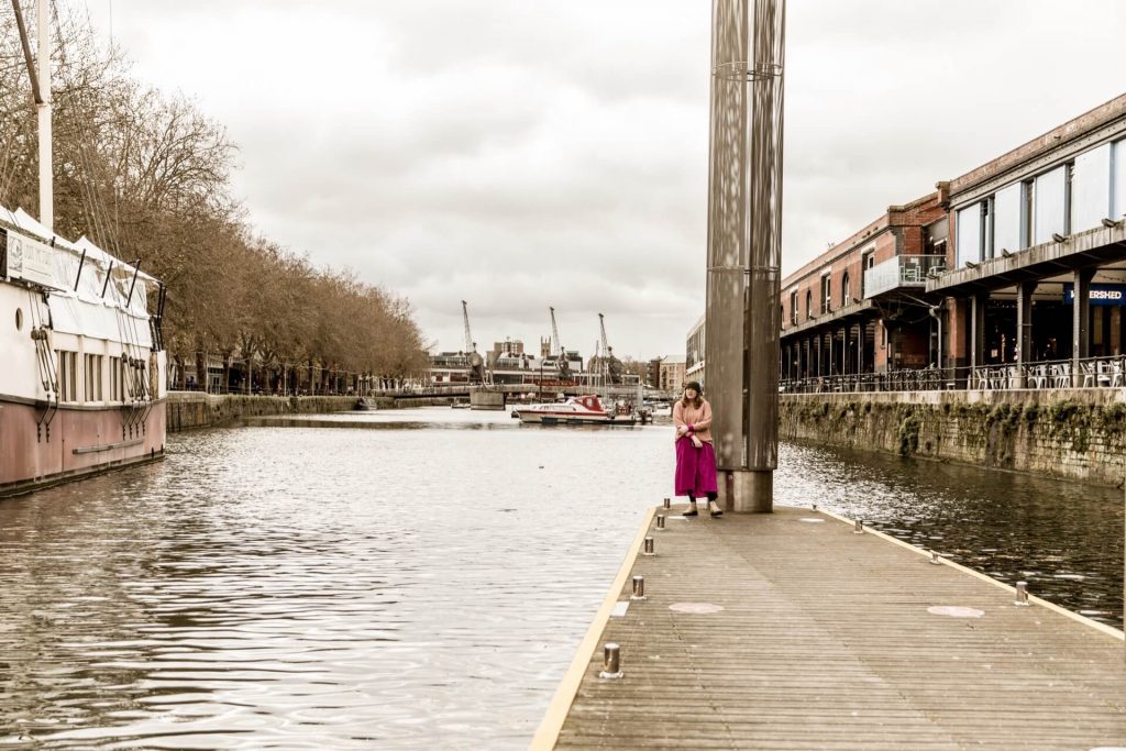Aime pondering how to make the mayor see that the WECA logo design competition is bad for creatives, while standing on a Bristol dock wearing a bright pink dress