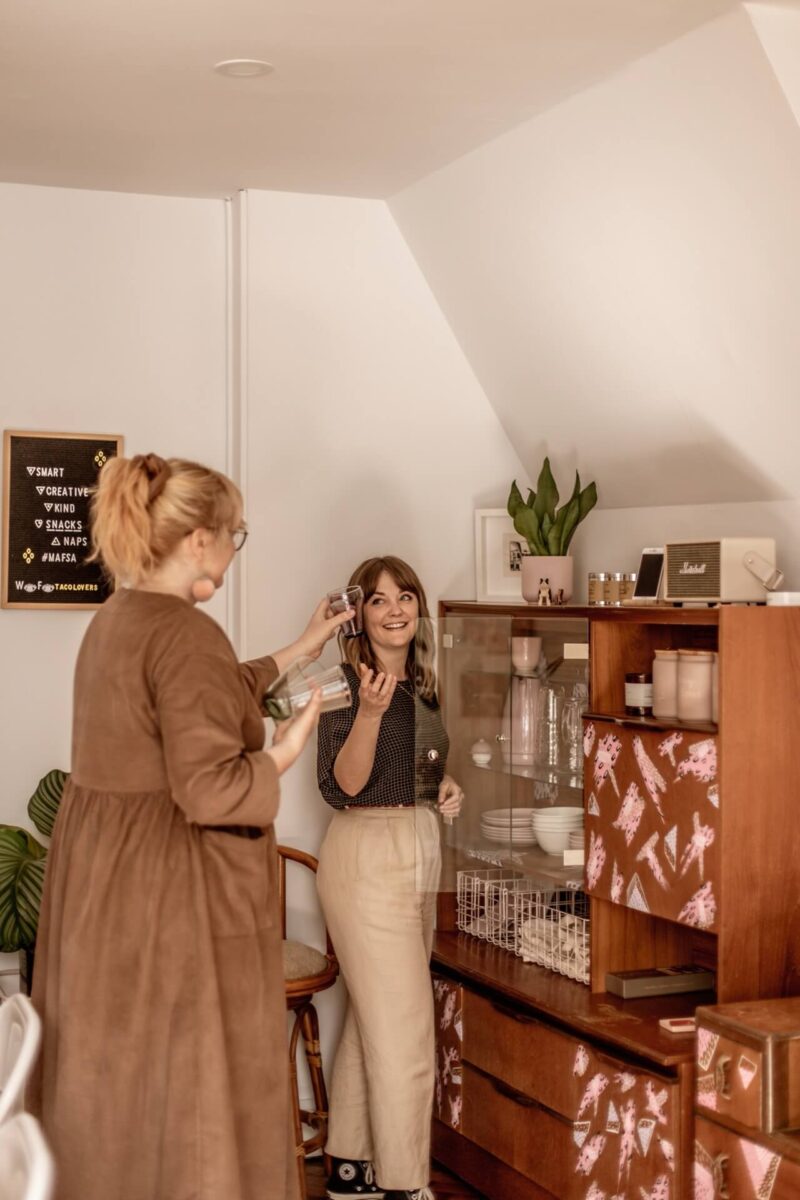 Content producer Lyzi wears a black top and beige trousers, and is smiling while handing drinking glasses to Studio Cotton founder Aime (wearing a brown dress) from the vintage cabinet, hand-painted by Emotional Waterfall Art.