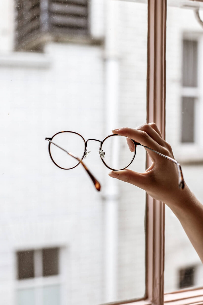 Hand holding a pair of delicate round reading glasses in front of a window