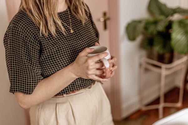 Close up shot from the neck down of copywriter Lyzi wearing a black grid-print top and beige trousers, holding a pink and white mug. In the background is a pink door, and a plant on a pink plant stand.