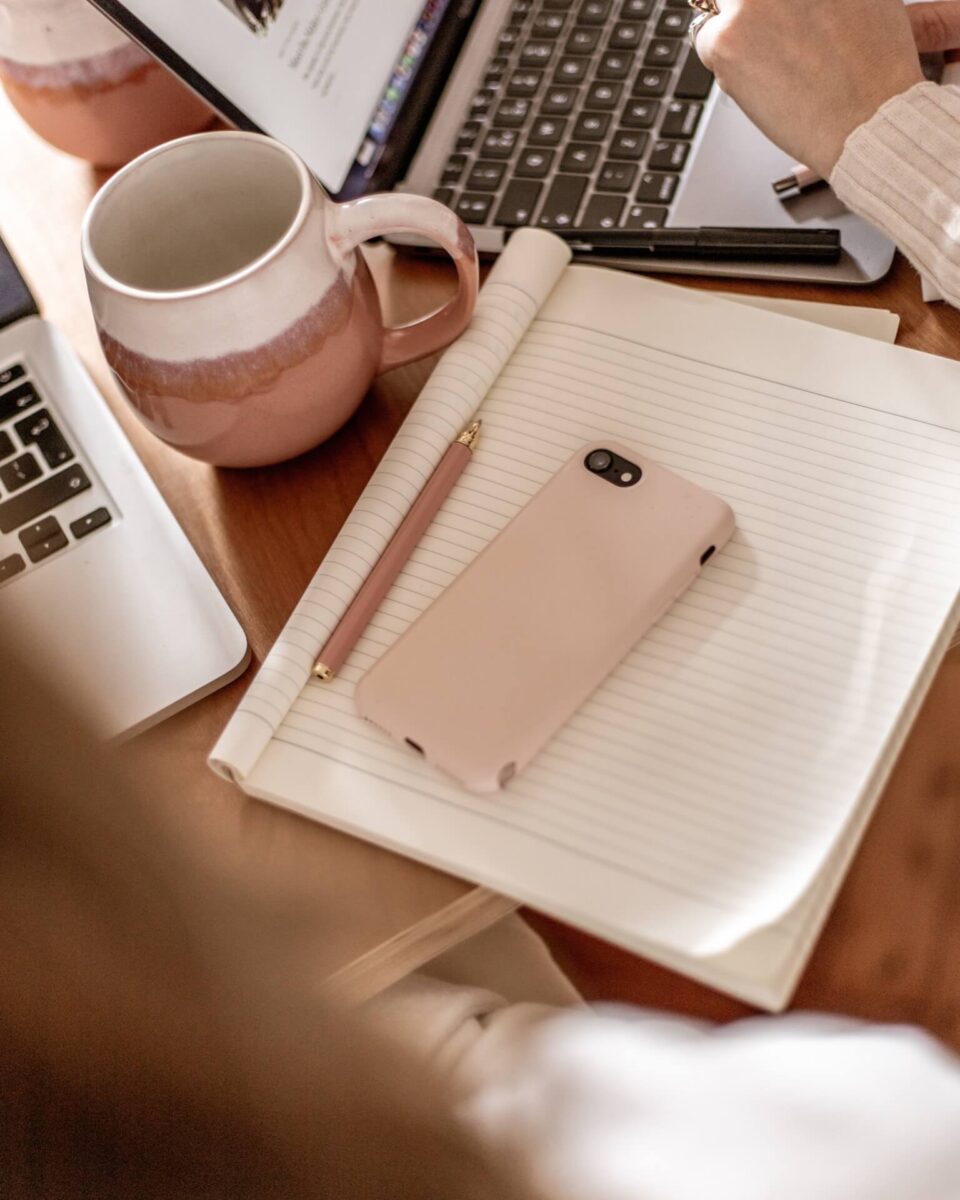 Shot of a phone with a pink case on top of an open notebook with pink pen, next to 2 laptops and pink and white mugs