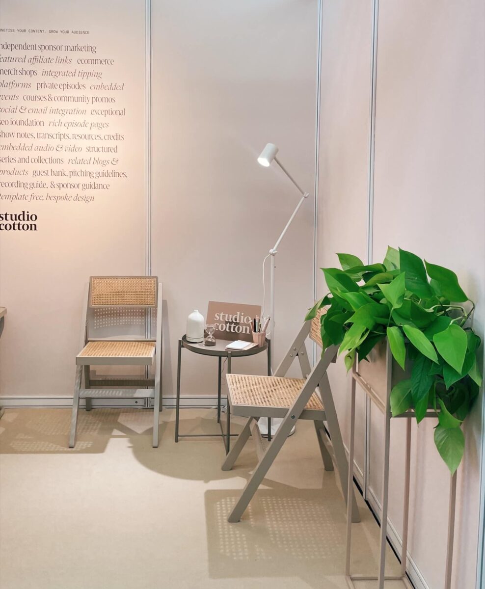 The Studio Cotton stand at The Podcast Show with a small side table and chairs, leafy plant, and text on the wall explaining our podcast website service